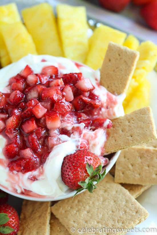 Bowl of strawberry fruit dip with a strawberry jam mixture on top of dip and a whole strawberry dipped into side with graham crackers