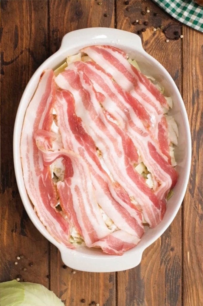 Raw bacon on top of cabbage casserole in white oval baking dish