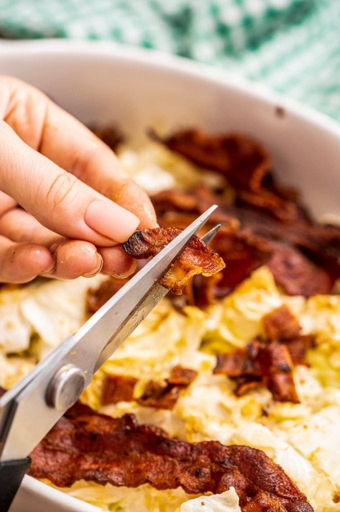 Kitchen shears cutting cooked bacon with cabbage casserole in background