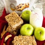 Two stacks of chewy apple cinnamon granola bars on a red kitchen towel with three green apples, a glass of milk, cinnamon sticks, dried apple slices, and a bag of truvia brown sugar