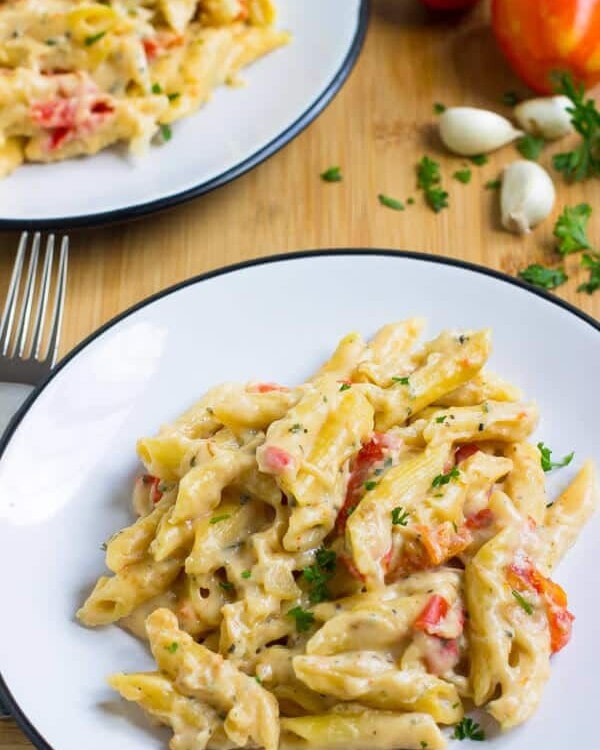 Two white plates with garlic and herb penne pasta on them sitting on a wood board with garlic, fresh parsley, and tomatoes