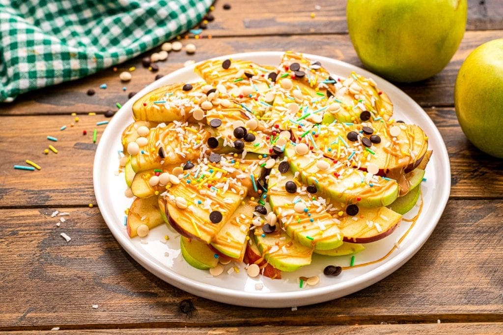 White plate with thinly sliced apples drizzled with peanut butter, chocolate chips, sprinkles and coconut.