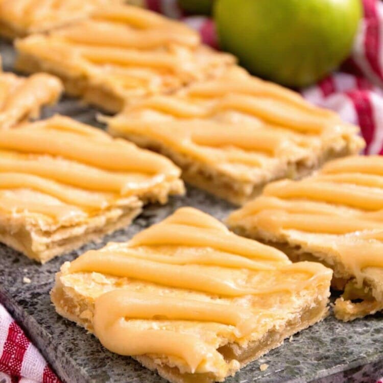 Eight apple pie bars on a stone tray sitting on a white and red kitchen towel along with two green apples
