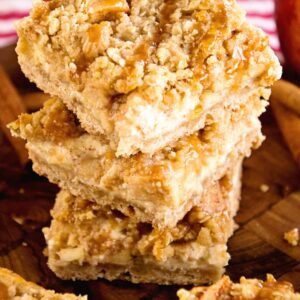 A stack of three caramel apple cheesecake bars on a wood board next to more bars and some apples