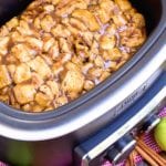 A crock pot of caramel apple rolls next to cinnamon sticks and green apples