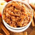 Two white bowls stacked with the top full of light crock pot applesauce sitting on a wood board next to cinnamon sticks and apples