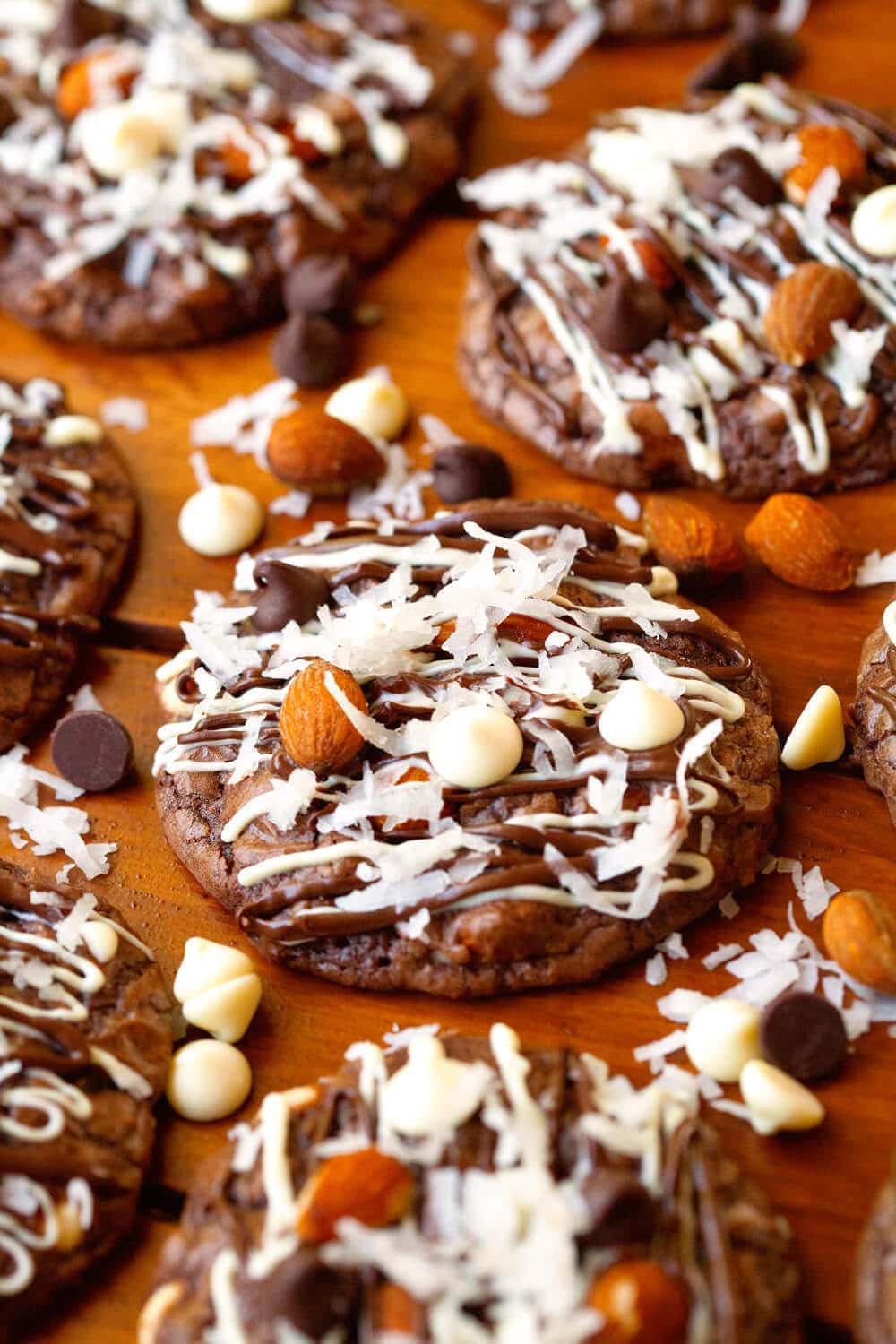 Almond Joy Cookies on wooden background and chocolate chips, almonds and white chocolate chips next to them.