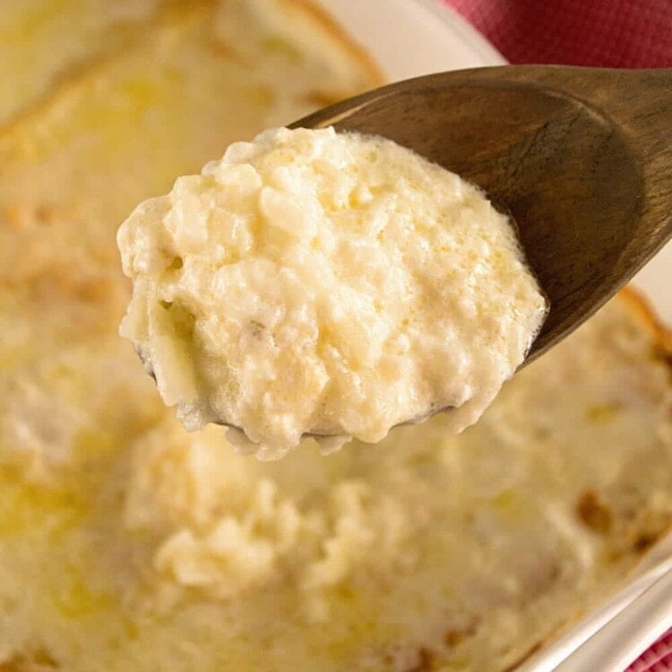 A wooden spoon with a scoop of hashbrown potatoes on it being held over a white casserole dish of hashbrown potatoes
