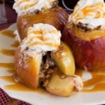 Three light crock pot baked apples with whipped cream and caramel sauce on a white plate sitting on a red kitchen towel with cinnamon sticks