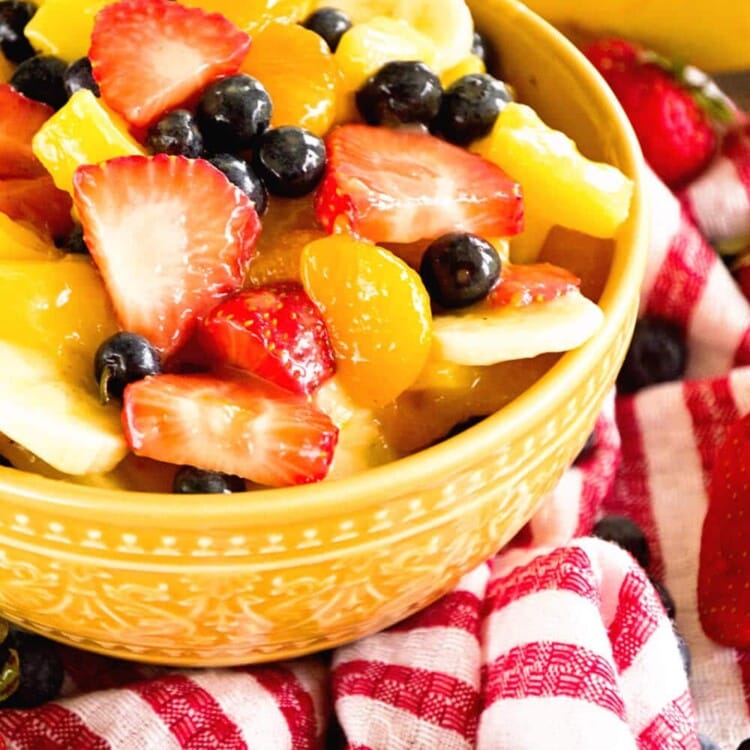A yellow bowl of sunshine fruit salad on a red and white striped kitchen towel along with strawberries, blueberries, and bananas