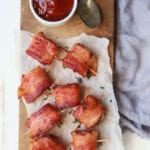 Bacon wrapped water chestnuts with toothpicks through them sitting on a piece of parchment paper next to a white bowl of sweet dipping sauce and a spoon all on a wood board