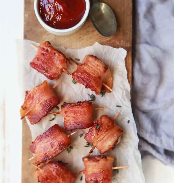 Bacon wrapped water chestnuts with toothpicks through them sitting on a piece of parchment paper next to a white bowl of sweet dipping sauce and a spoon all on a wood board