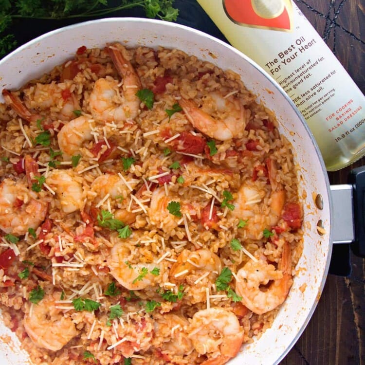Overhead image of light Italian shrimp and rice in a skillet on a wood table next to fresh parsley and a bottle of Thrive oil