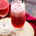 Bottle pouring bubbly into a champagne glass next to another glass of strawberry bellini on a wood board