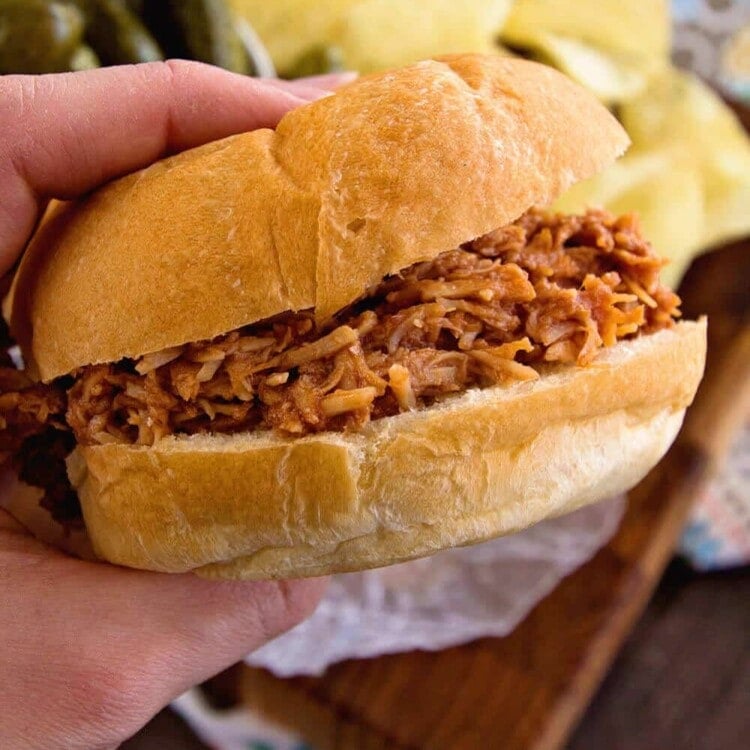 Hand holding a crock pot smokey bbq chicken sandwich over a board with pickles and potato chips on it