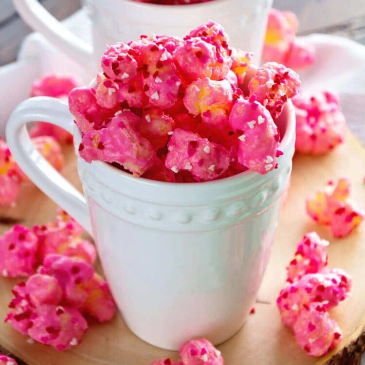 Two white mugs of puff corn valentines snack mix spilling out onto the wood platter they are on