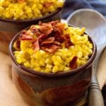 Two brown bowls of cheesy bacon ranch crock pot creamed corn with bacon on top sitting on a wood platter with a wooden spoon