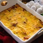 Ham and cheese overnight breakfast lasagna in a white casserole dish sitting on a red kitchen towel next to six eggs, an onion, and fresh parsley