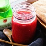 A glass mason jar of easy strawberry rhubarb jam on a navy blue kitchen towel with two small wood spoon, a jar of truvia, and a loaf of bread in a brown glass loaf pan