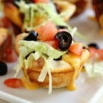 Taco muffin cups on a white square plate topped with cheese, lettuce, tomatoes, and olives