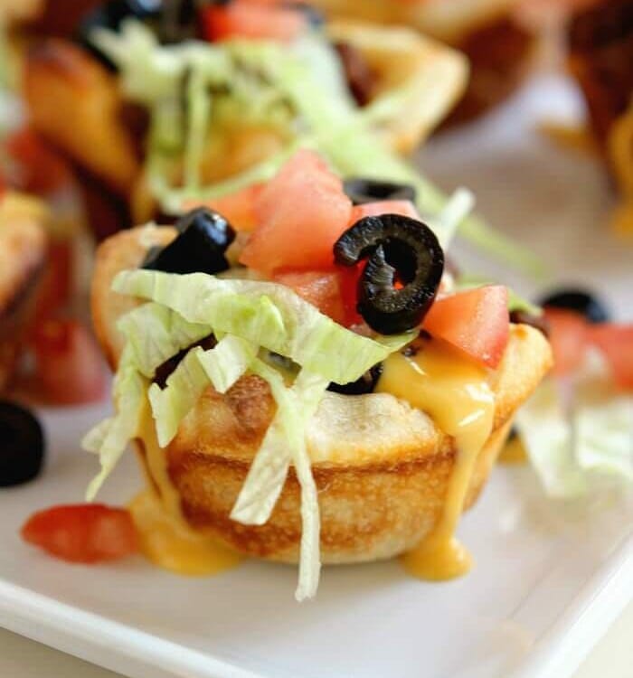 Taco muffin cups on a white square plate topped with cheese, lettuce, tomatoes, and olives