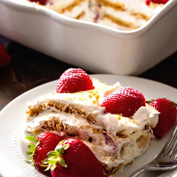 A piece of no bake strawberry cheesecake icebox cake on a white plate with a fork and two strawberries in front of a white baking dish containing the rest of the cake