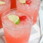 Three glasses of sparkling cherry limeade with a lime slice and a cherry in each one sitting on a white and gray chevron table cloth