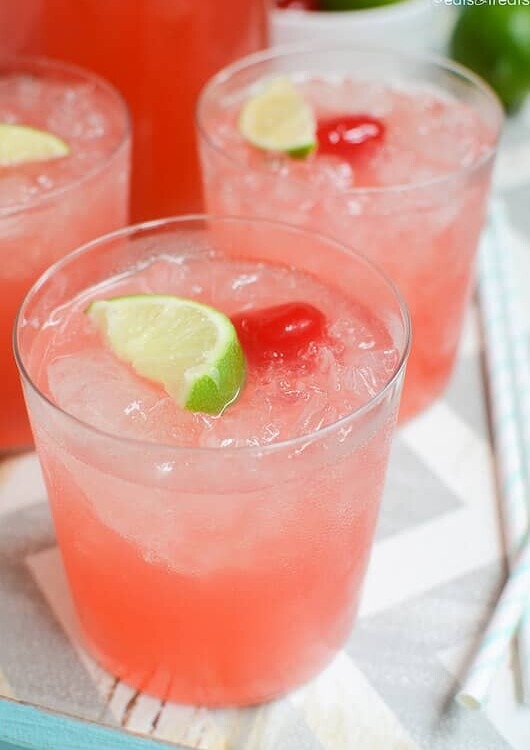 Three glasses of sparkling cherry limeade with a lime slice and a cherry in each one sitting on a white and gray chevron table cloth