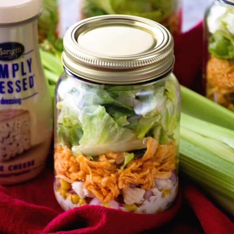 A mason jar of buffalo chicken salad on a red towel with celery and a jar of blue cheese dressing