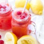Two clear glass mason jar mugs of raspberry lemonade sitting on a white counter with lemons and raspberries