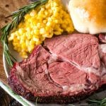 Slice of smoked prime rib with corn, a dinner roll, and rosemary on a plate