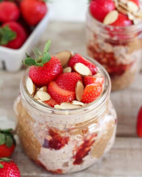 Two clear glass jars of strawberry almond overnight oats on a table with fresh strawberries on it