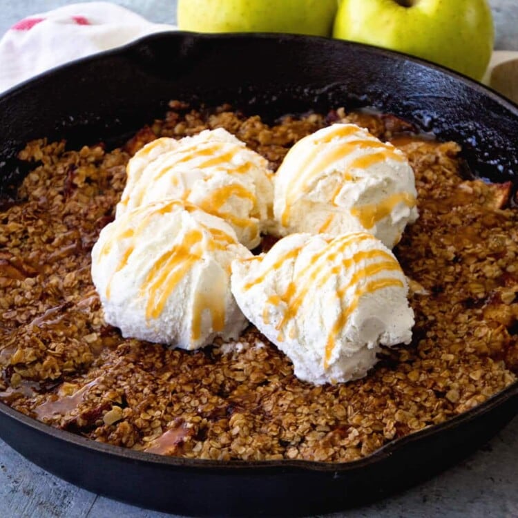 A cast iron skillet of grilled caramel apple crisp topped with four scoops of vanilla ice cream and caramel sauce