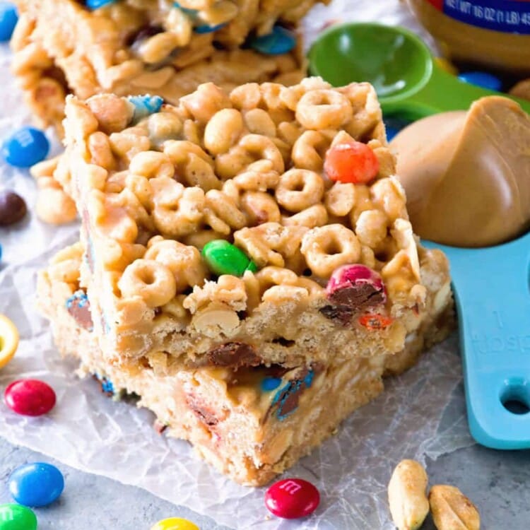 Two no bake peanut butter cereal bars stacked on a piece of wax paper along with m&m's, peanuts, measuring spoons, and a jar of peanut butter