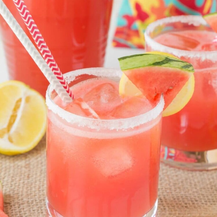 Two glasses with sugared rims of spiked watermelon lemonade and lemon and watermelon slices on the rim sitting on a burlap place mat