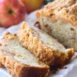 A partially sliced loaf of zucchini apple bread on a white tray in front of two apples and a zucchini