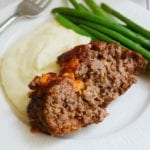 A slice of cheesy meatloaf on a white plate with mashed potatoes and green beans