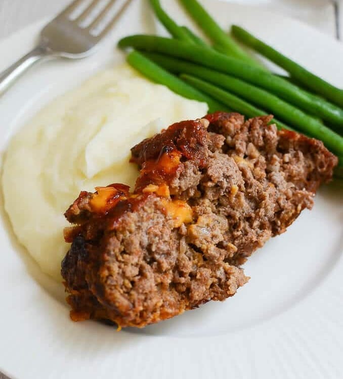 A slice of cheesy meatloaf on a white plate with mashed potatoes and green beans