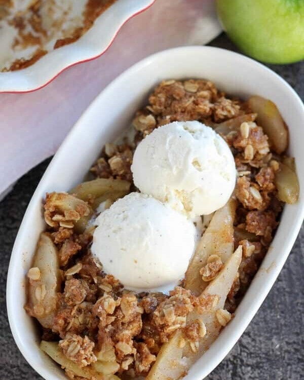 A white bowl of apple pear crisp topped with two scoops of vanilla ice cream