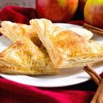 Three easy apple turnovers on a white plate sitting on a red kitchen towel with cinnamon sticks and apples