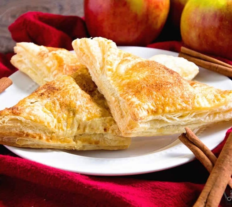 Three easy apple turnovers on a white plate sitting on a red kitchen towel with cinnamon sticks and apples