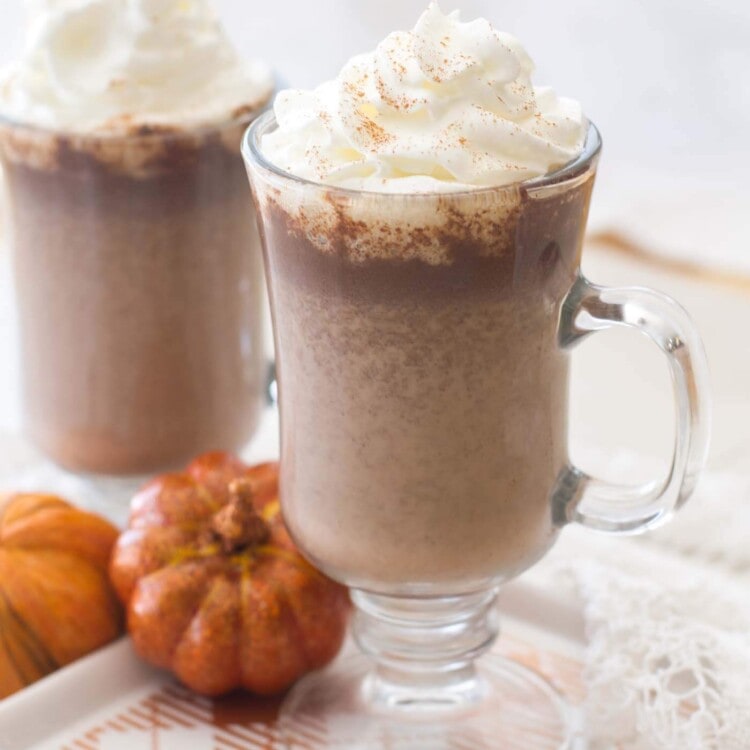 Two clear mugs of pumpkin hot chocolate topped with whipped cream sitting on a white and orange plate with decorative pumpkins