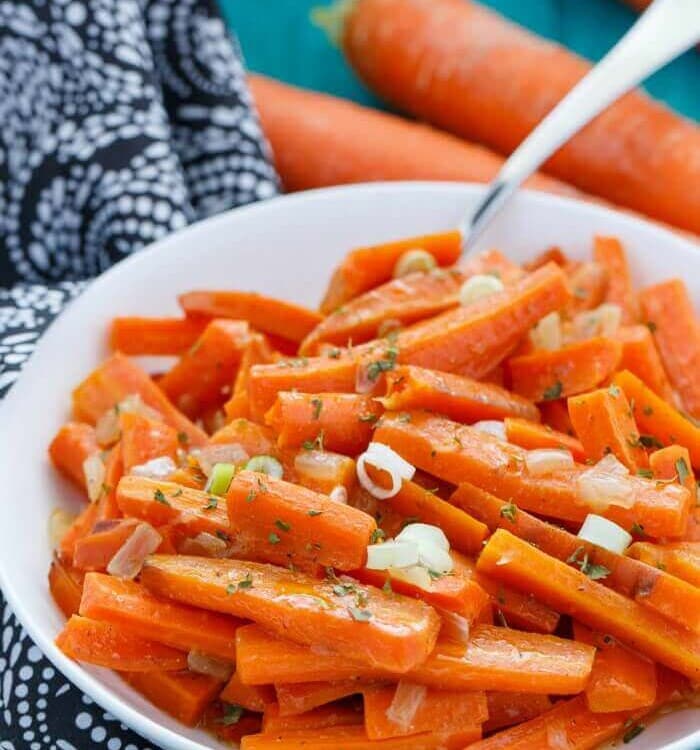 A white bowl of slow cooker ranch carrots on a blue table with a cloth napkin and three carrots