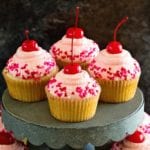 Four cherry almond cupcakes with heart sprinkles on a metal cake stand over more cupcakes