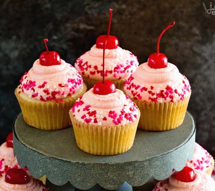 Four cherry almond cupcakes with heart sprinkles on a metal cake stand over more cupcakes