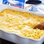 Creamy corn macaroni and cheese casserole in a white baking dish with a wood spoon in it and two bags of shredded cheese and a butter dish behind it