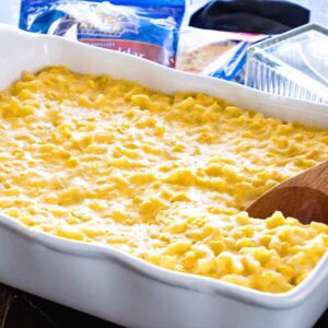 Macaroni de milho cremoso e caçarola de queijo numa assadeira branca com uma colher de pau e duas sacos de queijo desfiado e um prato de manteiga atrás dele