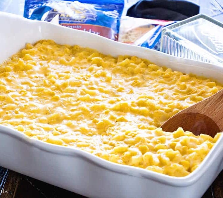 Creamy corn macaroni and cheese casserole in a white baking dish with a wood spoon in it and two bags of shredded cheese and a butter dish behind it