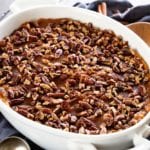 A white oval baking dish of light sweet potato casserole on a navy blue kitchen towel with cinnamon sticks, pecans, and a wood spoon