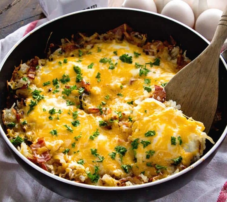 Cheesy bacon and egg hash brown skillet in a pan with a wooden spatula on a white towel